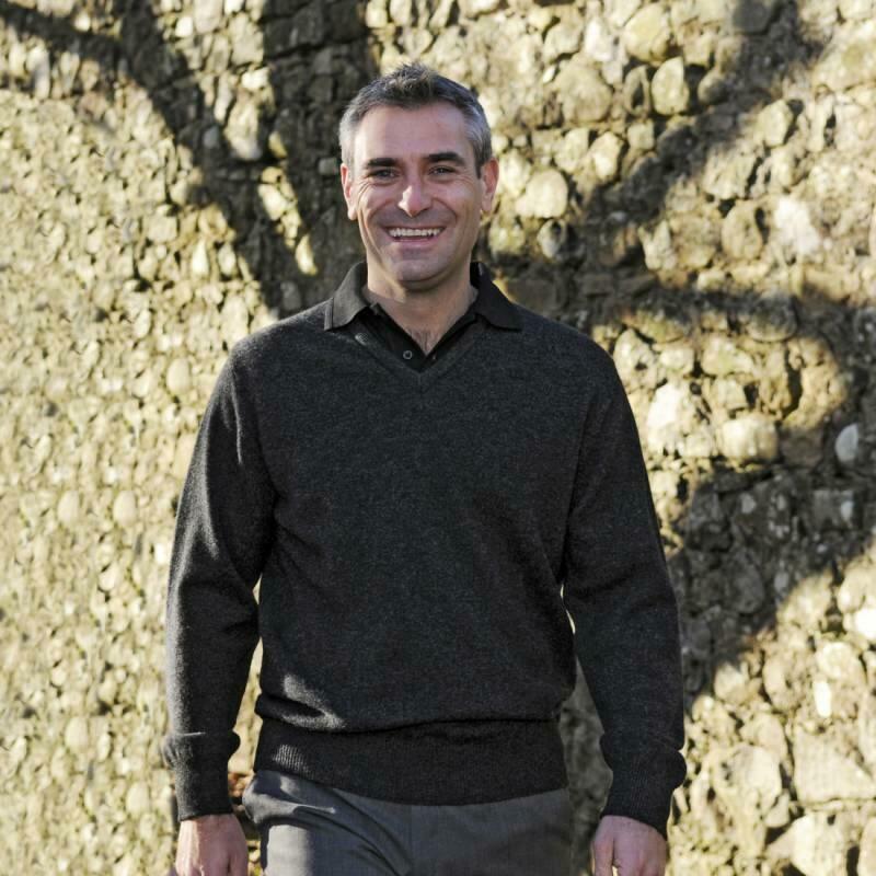 A smiling man with short gray hair is walking outdoors on a sunny day. He is wearing a dark gray Balmoral Lambswool sweater and pants, and the background shows a stone wall with shadows of tree branches cast on it.