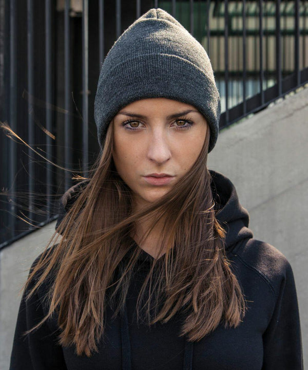 A person with long brown hair wears a gray beanie and a black Build Your Brand BY001 hoodie, standing in front of a metal fence. The wind is blowing strands of hair across their face. They have a serious expression and are looking directly at the camera.