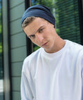 A young person with short hair, wearing a navy blue beanie and a white Build Your Brand BY002 sweatshirt, stands outside in front of modern building windows. They have a serious expression and are looking slightly downward toward the camera.