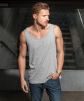 A man with short, styled hair and a beard stands in front of a stairwell, wearing a grey Build Your Brand BY003 sleeveless shirt and blue jeans. He has one hand in his pocket and wears a watch on his left wrist, looking off to the side.