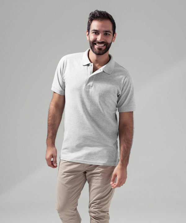 A man with dark hair and a beard smiles while standing against a plain grey background. He is wearing a light grey BY008 polo shirt from Build Your Brand and beige pants. He has one hand relaxed by his side and the other slightly raised.