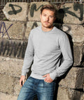 A man with short hair and a beard leans against a stone wall covered in graffiti. He is wearing a light grey, long-sleeve shirt and blue jeans from Build Your Brand's BY010 collection. The expression on his face is neutral, and he has his hands in his pockets.