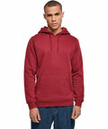 A person wearing a solid red Build Your Brand BY011 hoodie and blue jeans stands facing the camera against a plain white background. The hoodie has a front pouch pocket and drawstrings on the hood. The person has a neutral expression and short hair.