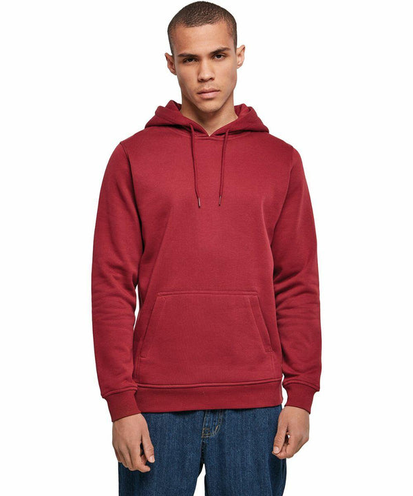 A person wearing a solid red Build Your Brand BY011 hoodie and blue jeans stands facing the camera against a plain white background. The hoodie has a front pouch pocket and drawstrings on the hood. The person has a neutral expression and short hair.
