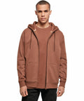 A person stands facing the camera, wearing a brown Build Your Brand BY012 zip-up hoodie over a matching brown t-shirt. The hoodie has drawstrings and two large front pockets. The person has short hair and a neutral expression. The background is plain and white.