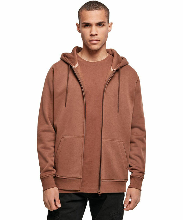 A person stands facing the camera, wearing a brown Build Your Brand BY012 zip-up hoodie over a matching brown t-shirt. The hoodie has drawstrings and two large front pockets. The person has short hair and a neutral expression. The background is plain and white.