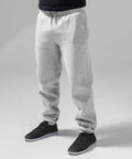 A person stands wearing BY014 grey sweatpants by Build Your Brand and black casual shoes. The background is a solid grey, highlighting the comfortable and relaxed nature of the outfit. Only the lower half of the person's body is visible.