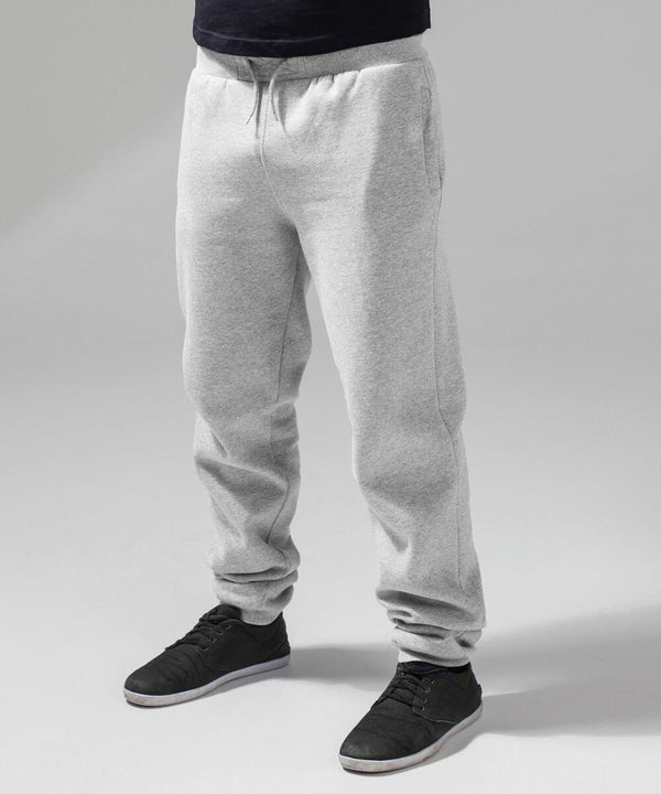A person stands wearing BY014 grey sweatpants by Build Your Brand and black casual shoes. The background is a solid grey, highlighting the comfortable and relaxed nature of the outfit. Only the lower half of the person's body is visible.