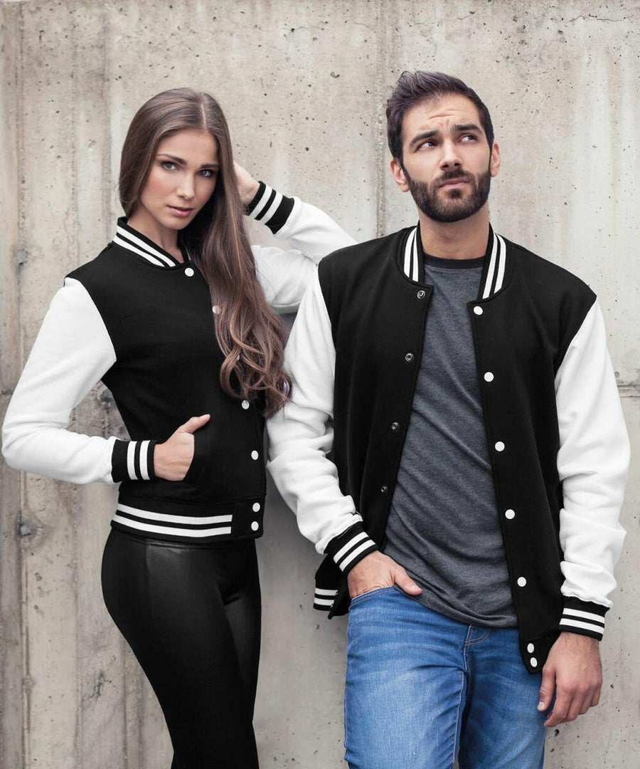 Two models stand in front of a concrete wall wearing black and white varsity jackets. The woman on the left has long hair and is posing with one hand on her head and the other in her pocket. The man on the right has a beard, a hand in his pocket, and looks away. They are showcasing BY015 by Build Your Brand.