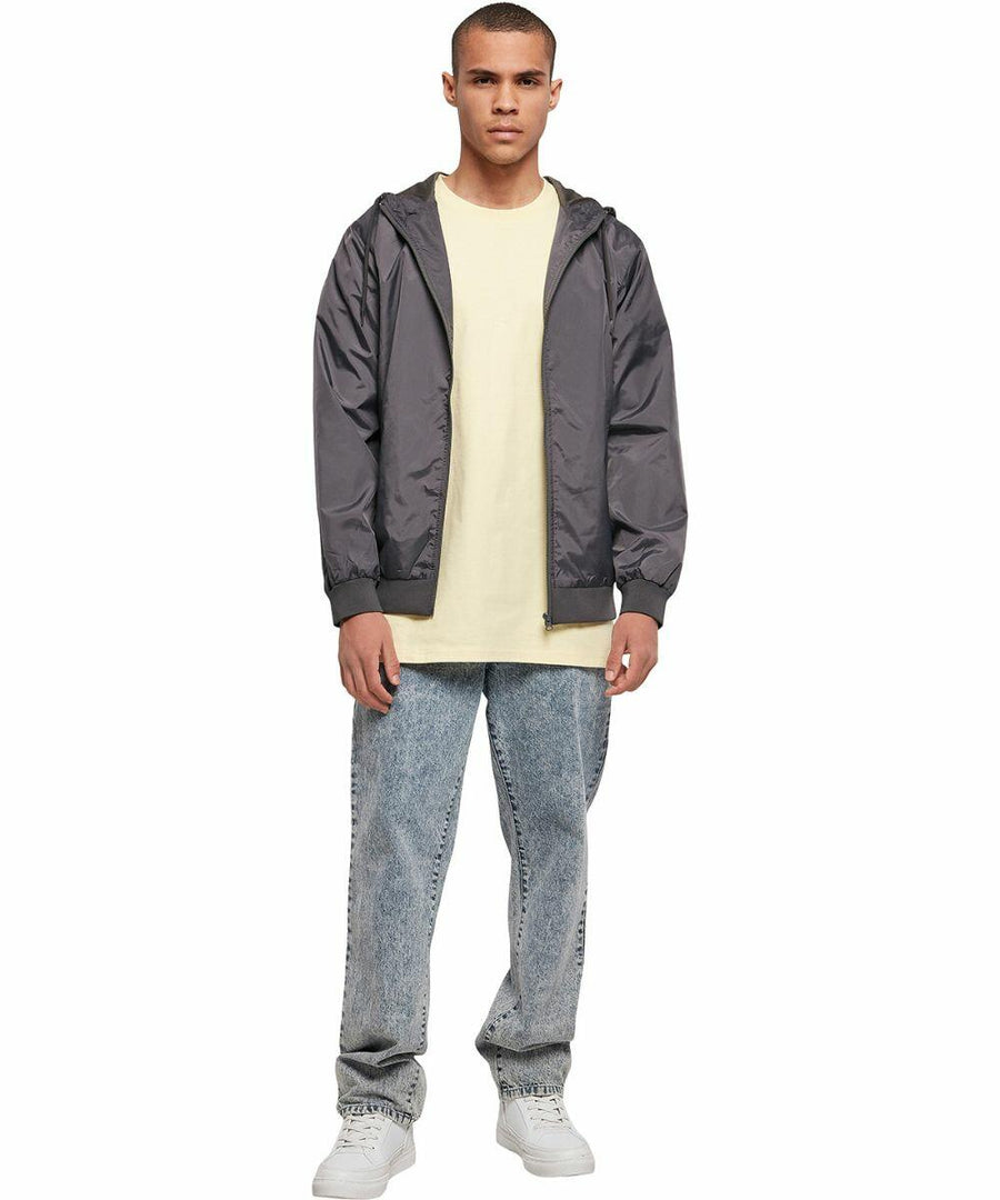 A person stands facing the camera wearing a gray Build Your Brand BY016 zip-up jacket over a light yellow t-shirt, light blue jeans, and white sneakers. The background is plain white.