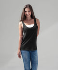 A person with long brunette hair is smiling while standing against a gray background. They are wearing a black sleeveless BY019 top by Build Your Brand over a white undershirt and blue jeans. Their left arm is positioned behind their back.