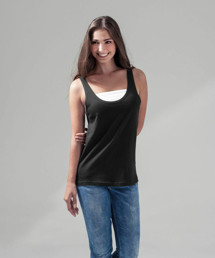 A person with long brunette hair is smiling while standing against a gray background. They are wearing a black sleeveless BY019 top by Build Your Brand over a white undershirt and blue jeans. Their left arm is positioned behind their back.