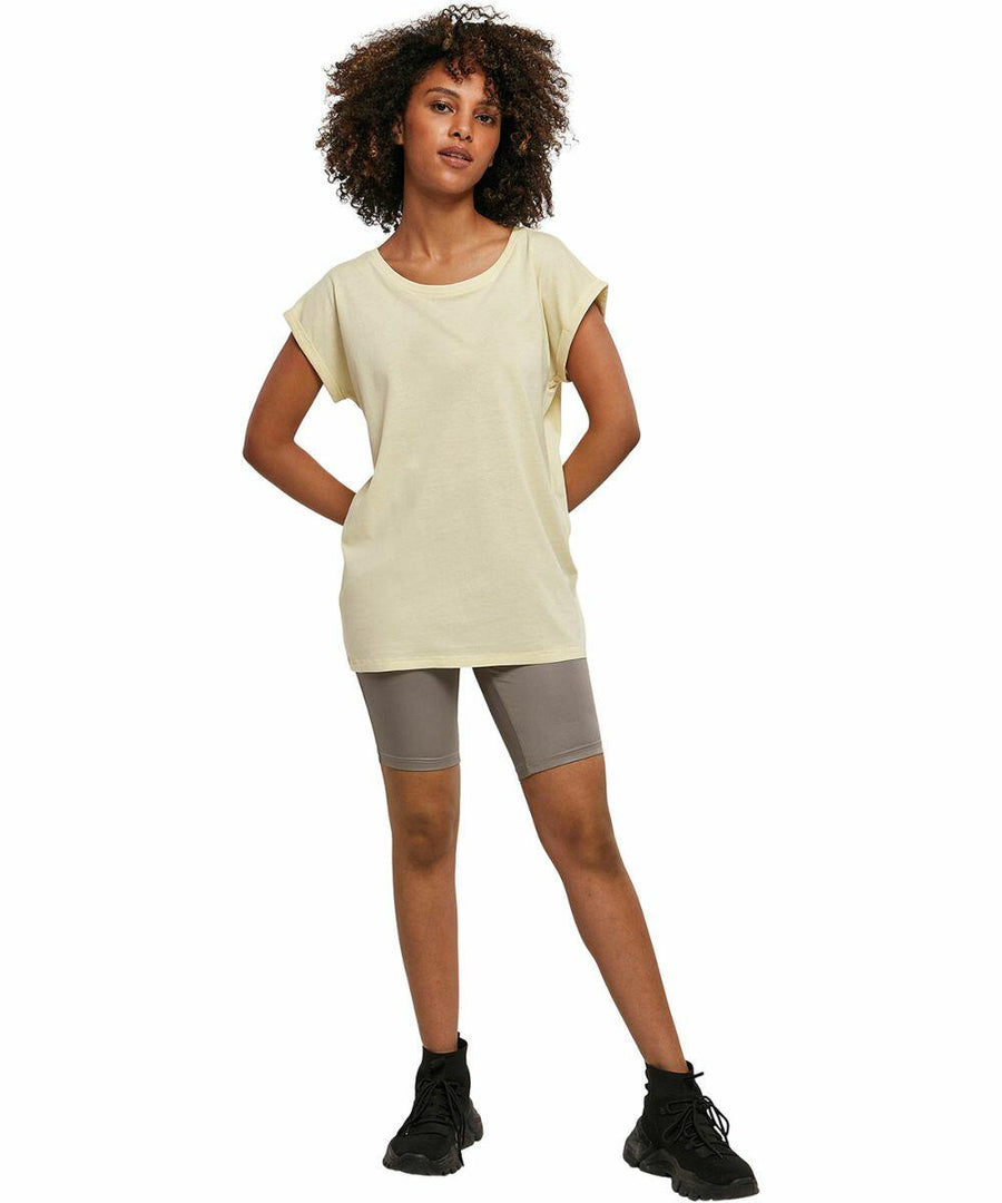 A person with curly hair stands confidently with hands on hips. They are wearing a Build Your Brand BY021 light yellow t-shirt, gray shorts, and black sneakers. The background is plain white.