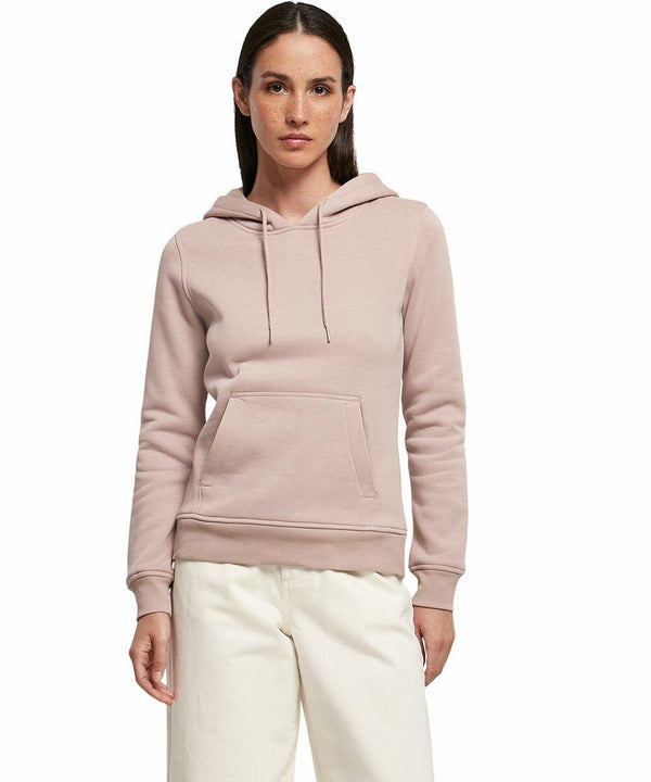 A person with long dark hair is wearing a light pink Build Your Brand BY026 hoodie with a front pocket and white drawstrings. They are also wearing white pants and are standing against a white background. They have a neutral expression and are facing forward.