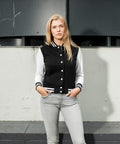 A woman with long blonde hair stands against a gray wall. She is wearing a black BY027 varsity jacket by Build Your Brand with white sleeves and gray jeans. Her hands are in her pockets, and she has a neutral expression on her face.