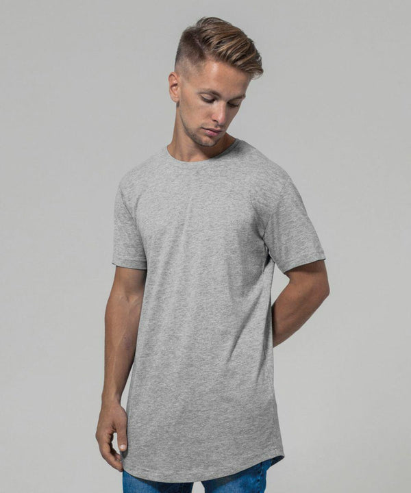 A person with short, styled hair and a trimmed beard is wearing a fitted Build Your Brand BY028 light gray T-shirt and blue jeans. The person is looking down and has one hand behind their back against a plain gray background.