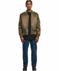 A man stands facing the camera wearing a BY030 bomber jacket by Build Your Brand over a black shirt, blue jeans, and black shoes. He has short, braided hair and a neutral expression. The background is plain white.