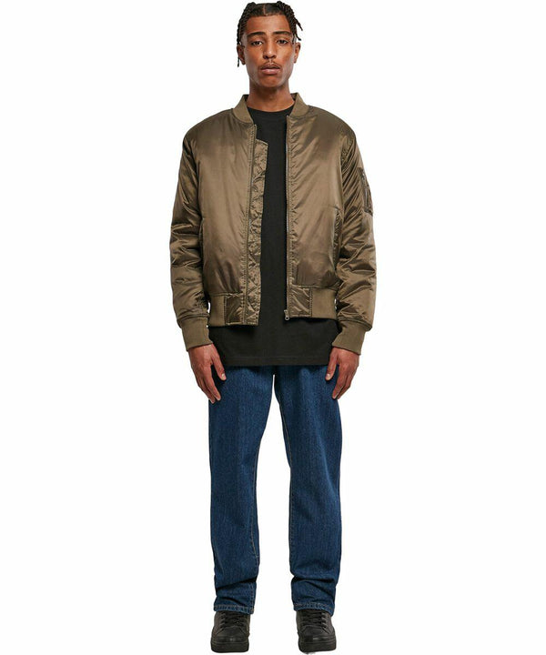 A man stands facing the camera wearing a BY030 bomber jacket by Build Your Brand over a black shirt, blue jeans, and black shoes. He has short, braided hair and a neutral expression. The background is plain white.