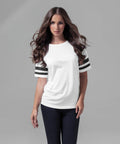 A woman with long, wavy brown hair is wearing a white Build Your Brand BY033 t-shirt with black and white striped sleeves and black pants. She is standing against a light gray background and looking directly at the camera.