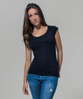 A person with long brown hair, wearing a black short-sleeved BY035 top from Build Your Brand and blue jeans, poses against a plain grey background. Their left hand rests casually on their hip while their right arm hangs by their side.
