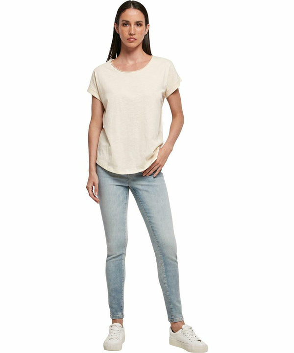 A person with long hair is wearing a short-sleeved, white BY036 t-shirt by Build Your Brand, light blue skinny jeans, and white sneakers. They are standing against a plain white background.