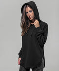 A person with long brown hair is wearing a black Build Your Brand BY037 hoodie with the hood up and dark-patterned pants, standing against a plain gray background. They have a serious expression and one hand is holding the edge of the hoodie hood.