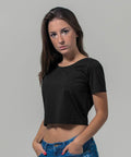 A person with long, dark hair is wearing a black Build Your Brand BY042 short-sleeved crop top and blue jeans. Their hands are in their pockets, and they are looking directly at the camera with a neutral expression. The background is plain gray.