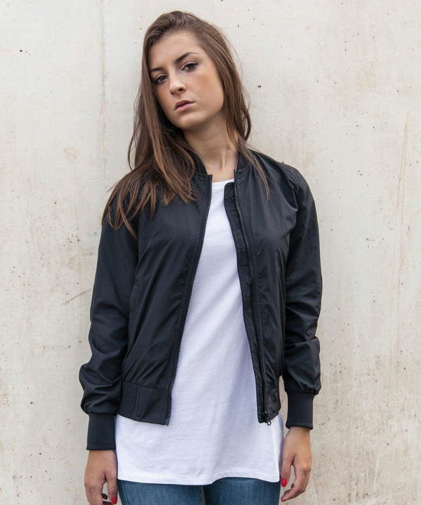 A woman with long brown hair stands against a plain, weathered wall. She wears a white T-shirt under a black Build Your Brand BY044 zip-up jacket and blue jeans. Her expression is serious, and her hands hang at her sides.