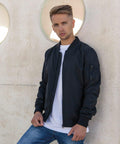 A person with short, styled hair stands against a concrete wall with circular cut-outs. They are wearing a black bomber jacket over a white t-shirt and blue jeans from Build Your Brand's BY045 line, looking directly at the camera with a serious expression.