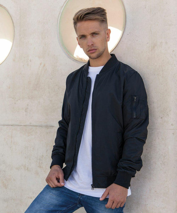 A person with short, styled hair stands against a concrete wall with circular cut-outs. They are wearing a black bomber jacket over a white t-shirt and blue jeans from Build Your Brand's BY045 line, looking directly at the camera with a serious expression.