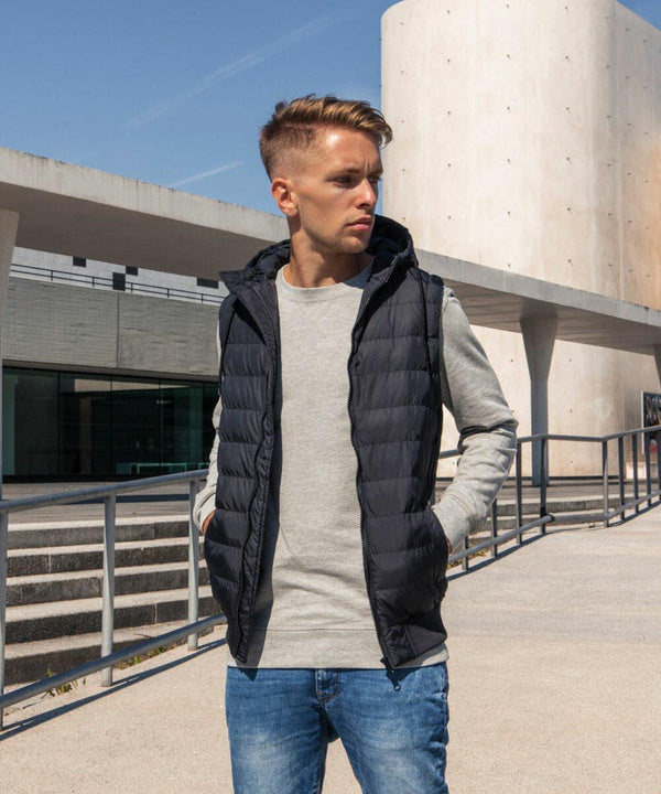 A man wearing a gray long-sleeve shirt and a black puffer vest from Build Your Brand, product name BY046, stands on a modern concrete patio with a large cylindrical building in the background. He is looking to the left with his hands in his vest pockets. The weather appears sunny and clear.