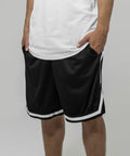 A person dressed in a white Build Your Brand BY047 T-shirt and black athletic shorts with white stripes is standing with hands in pockets. The image shows them from the shoulders down against a plain gray background.
