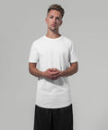 A man with short, styled hair is wearing a BY048 plain white t-shirt from Build Your Brand and black pants. He is standing against a gray background with his hands clasped together in front of him, looking directly at the camera.