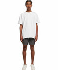 A person stands facing forward wearing a loose-fitting white BY050 T-shirt from Build Your Brand and gray shorts. They are also wearing white sneakers. The background is plain white.