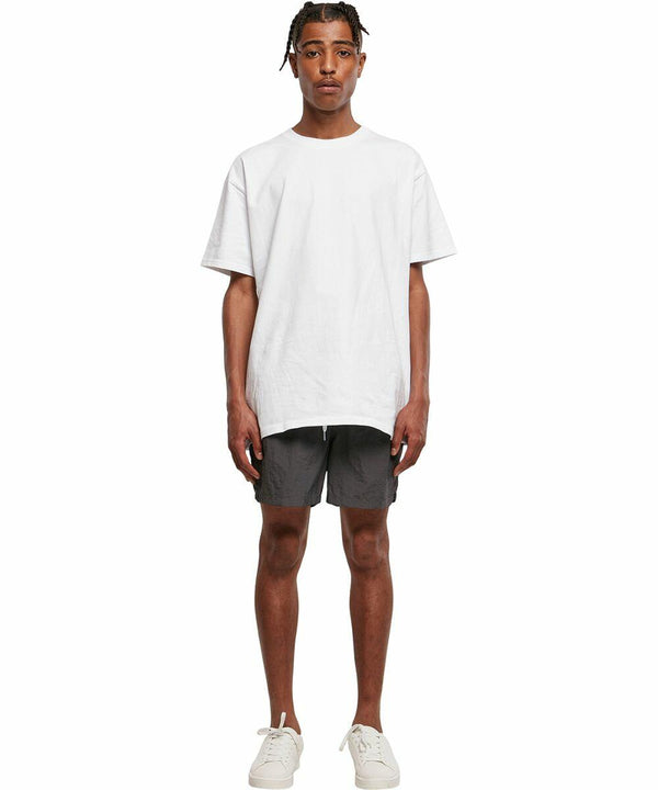 A person stands facing forward wearing a loose-fitting white BY050 T-shirt from Build Your Brand and gray shorts. They are also wearing white sneakers. The background is plain white.