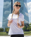 A person wearing sunglasses and a white Build Your Brand t-shirt with braided hair is standing outdoors. A modern glass skyscraper and lush greenery are visible in the background under a partly cloudy sky.