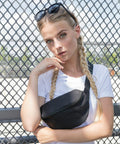 A young woman with long blond braids and sunglasses on her head stands in front of a chain-link fence. She wears a white Build Your Brand BY059 t-shirt and a black crossbody bag. She gazes into the camera with a pensive expression, her right hand lightly touching her chin.