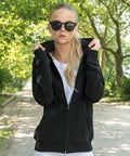 A blonde woman wearing round, dark sunglasses and a black Build Your Brand BY069 zip-up hoodie stands on a tree-lined path. She is pulling the hood slightly over her shoulders and looking directly at the camera. The background includes green trees and sunlight filtering through the leaves.