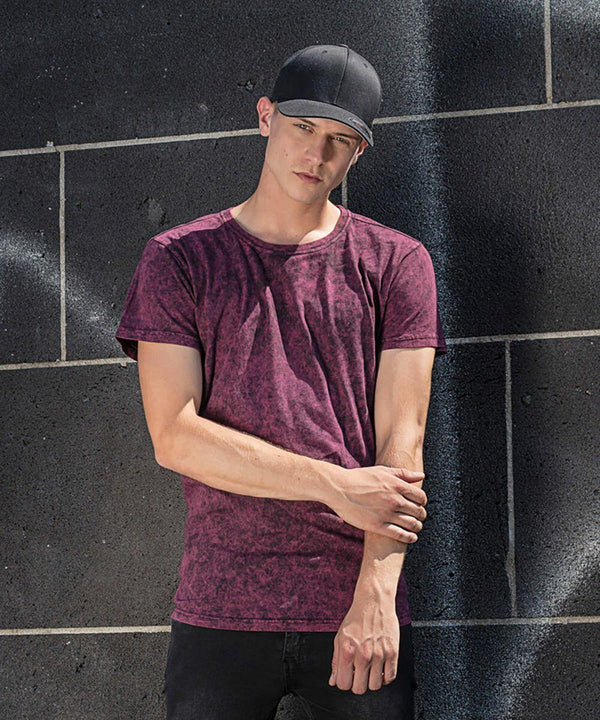 A young person wearing a black cap, a maroon tie-dye t-shirt, and black jeans from Build Your Brand stands against a black brick wall. They have their head slightly tilted down and their hands clasped together in front of them. The lighting casts shadows on the wall.