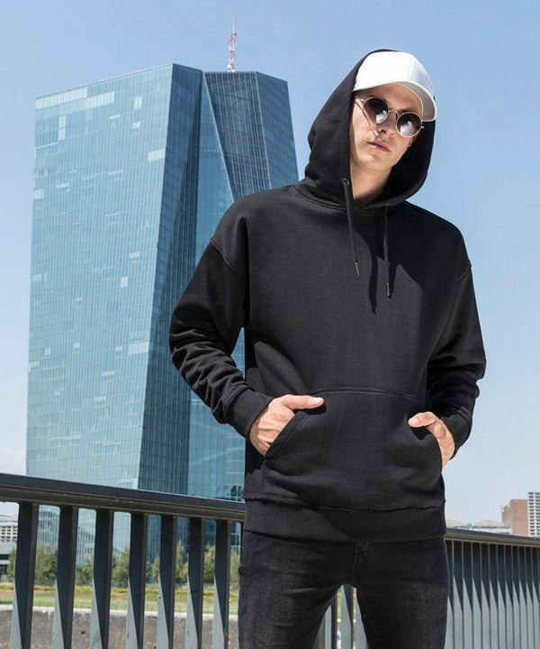 A person in a black BY074 hoodie by Build Your Brand, black pants, white cap, and sunglasses stands with hands in the hoodie pockets. They are posing in front of a modern glass building with an angular design on a clear day.