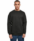 A person wearing a plain black Build Your Brand BY075 sweatshirt and blue jeans stands against a white background. The person has short hair and a neutral expression, with hands relaxed by their sides.