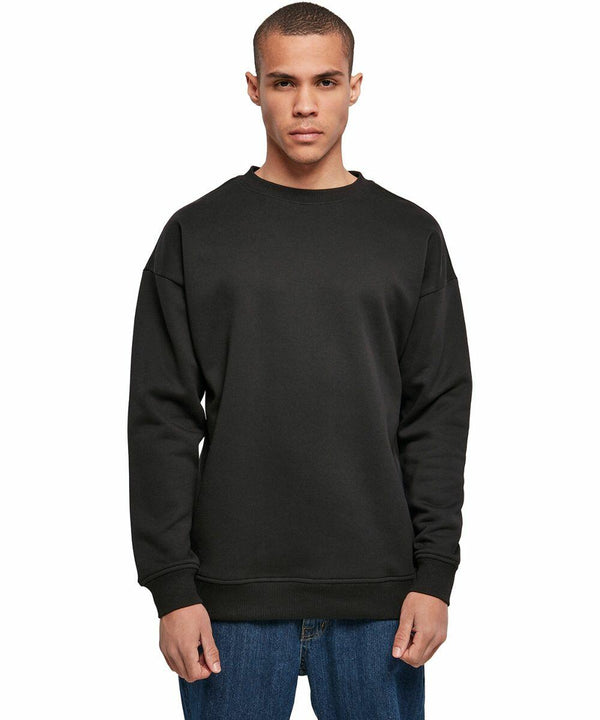 A person wearing a plain black Build Your Brand BY075 sweatshirt and blue jeans stands against a white background. The person has short hair and a neutral expression, with hands relaxed by their sides.