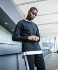 Young man in casual attire poses indoors, wearing a black long-sleeved shirt over a BY091 white shirt by Build Your Brand, black pants with white stripes, and bracelets on his right wrist. Modern architectural background.
