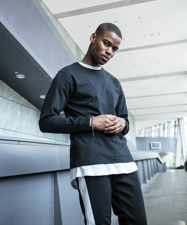 Young man in casual attire poses indoors, wearing a black long-sleeved shirt over a BY091 white shirt by Build Your Brand, black pants with white stripes, and bracelets on his right wrist. Modern architectural background.