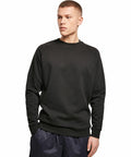 A person stands wearing a plain black BY094 long-sleeve sweatshirt from Build Your Brand and dark athletic pants against a white background.