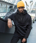 Person in a black hoodie and yellow beanie stands indoors, leaning against a railing with one arm. Modern, minimalistic architectural interior visible in the background. They are wearing the BY098 by Build Your Brand.