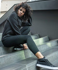 A person with curly hair sits on stairs wearing a black BY099 hoodie from Build Your Brand, black leggings, and black sneakers, looking directly at the camera.