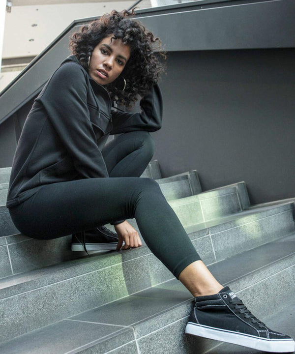 A person with curly hair sits on stairs wearing a black BY099 hoodie from Build Your Brand, black leggings, and black sneakers, looking directly at the camera.