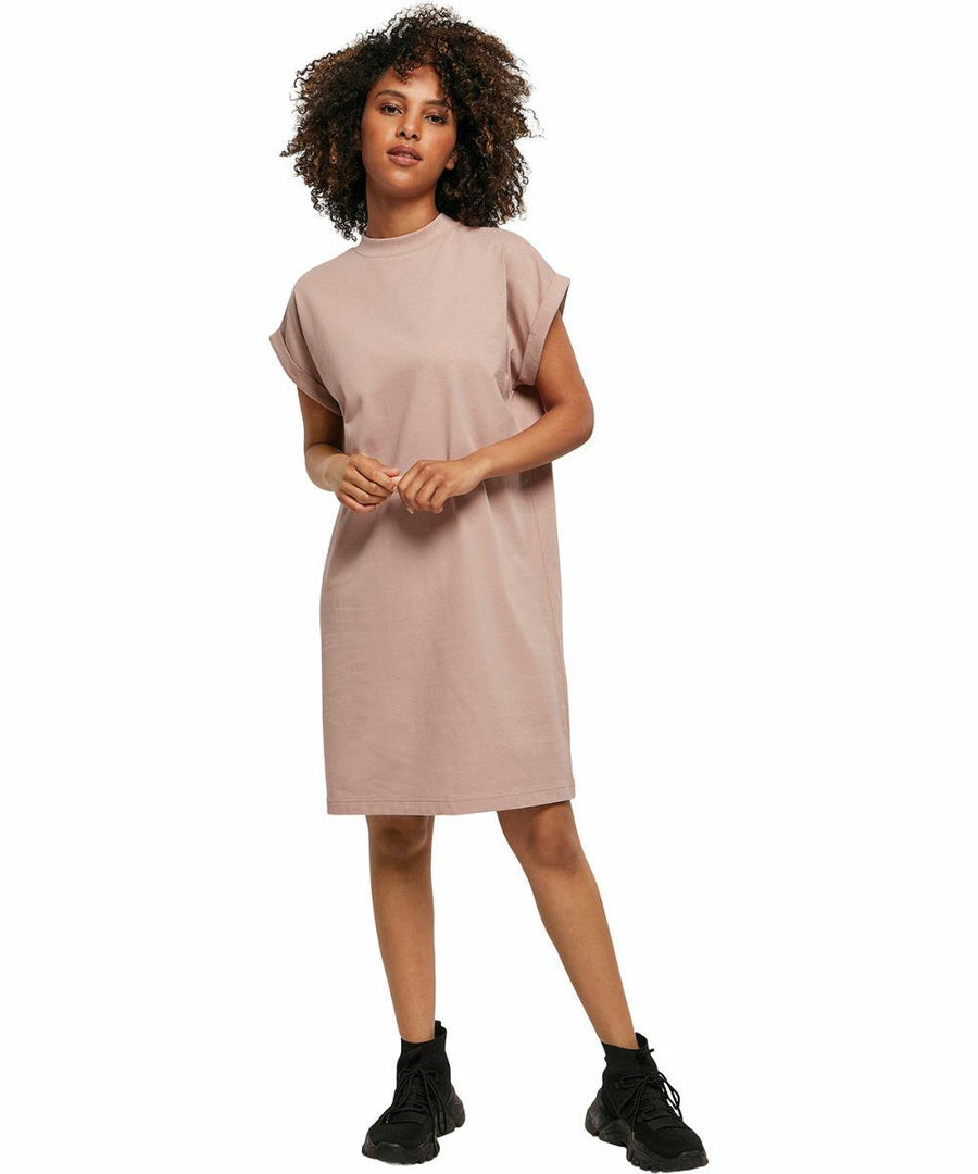 A woman with curly hair is wearing a knee-length, short-sleeved, loose-fitting pink dress and black BY101 sneakers by Build Your Brand. She is standing on a white background.