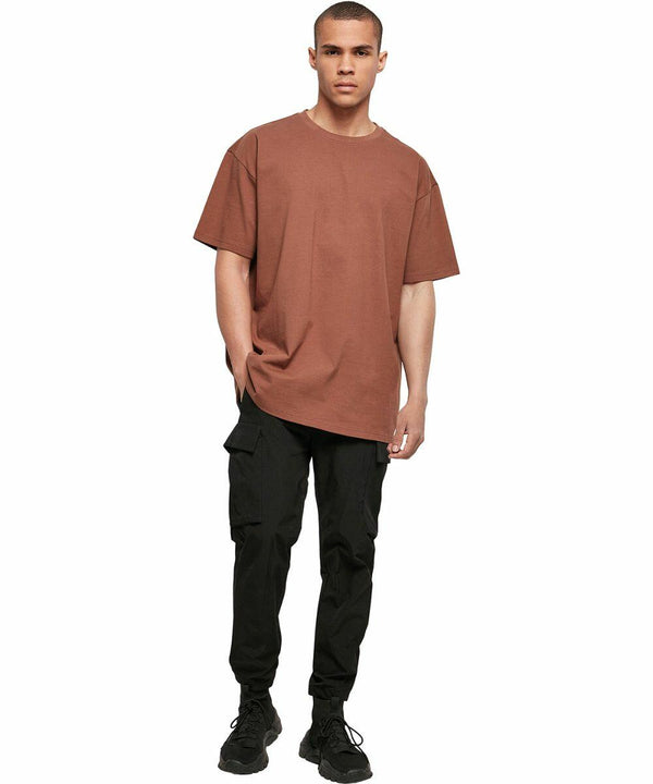 A person wearing a loose brown t-shirt, BY102 by Build Your Brand, black cargo pants, and black shoes is standing against a plain white background.